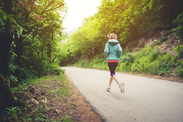 Fototapeta premium Young fitness blonde woman in headphones running at morning caucasian forest trail in sun light. side view from behind