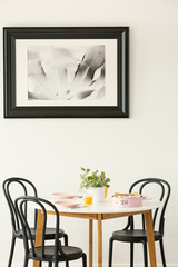 Large, black and white picture of a monstera leaf hanging on a white wall above a round dining table in bright apartment interior. Real photo
