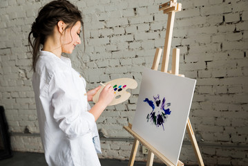 Portrait of beautiful brunette girl wearing in white shirt and jeans, drawing picture with oil paints. Smiling, pretty woman, young female artist dreaming about art and holding brush and palette.