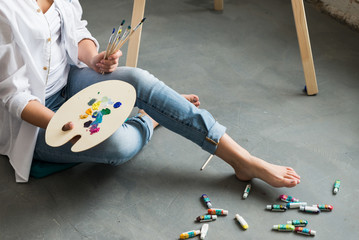 Crop of girl in white shirt and jeans, sitting at work place and creating, drawing picture with oil colorful paints. Pretty woman, female artist holding brush and palette. Concept of art.