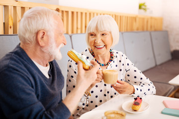 Delicious lunch. Joyful senior woman delighting coffee and senior man biting eclair