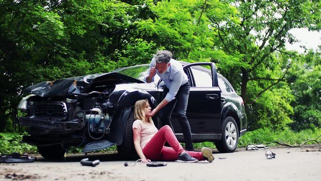 Injured woman by the car after an accident and a man making a phone call.