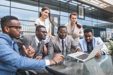 Group of happy diverse male and female business people team in formal gathered around laptop computer in bright office against the background of a glass building