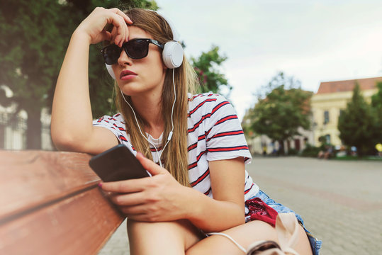 Girl with sunglasses sitting on a bench in the summer listening music over headphones
