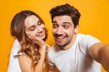 Portrait of excited smiling couple man and woman taking selfie photo together, isolated over yellow background