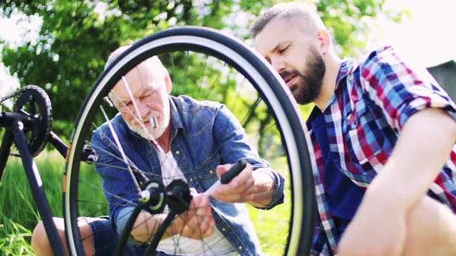 An adult hipster son and senior father repairing bicycle outside on a sunny day.