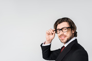 side view of smiling businessman in stylish suit and eyeglasses looking at camera isolated on grey