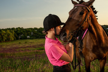 Beautiful smiling girl jockey stand next to her brown horse wearing special uniform on a sky and...