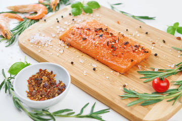 Salmon fillet lies on a wooden bamboo board on a white background. Prepared ingredients for cooking fish, rosemary, basil, shrimp, cherry tomatoes, lemon, spices, large Hawaiian salt