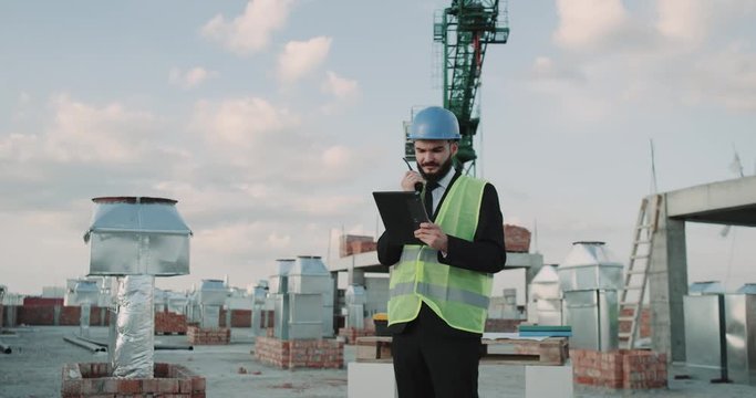 On rooftop at construction site businessman wearing a safety helmet and high visibility vest speaking using a black radio.