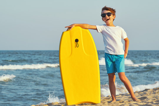 A happy boy in sunglasses in swimming trunks stands on the beach on golden sand and holds a swimming board, boogie board of yellow color against the blue sky and the sea with small waves in sunny sun