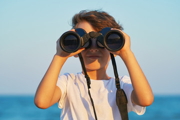 A happy boy holds a large pair of binoculars and stares into the distance on a blue sky against the blue sea and the blue sunset of a sunny day