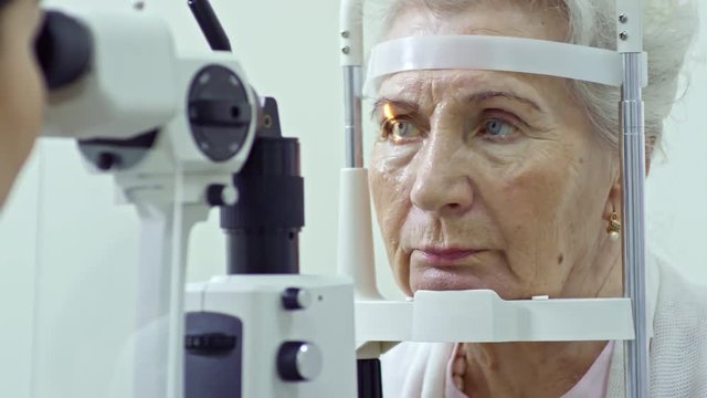Tilt up of elderly woman looking at female optometrist while she examining her eye with slit lamp