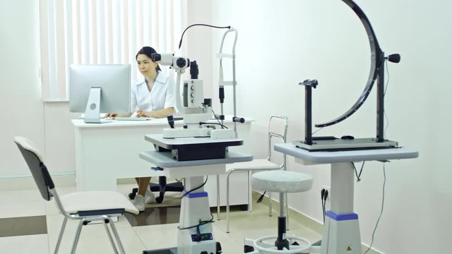 Tracking shot of female doctor in uniform sitting at desk and working on computer at her office in ophthalmology clinic