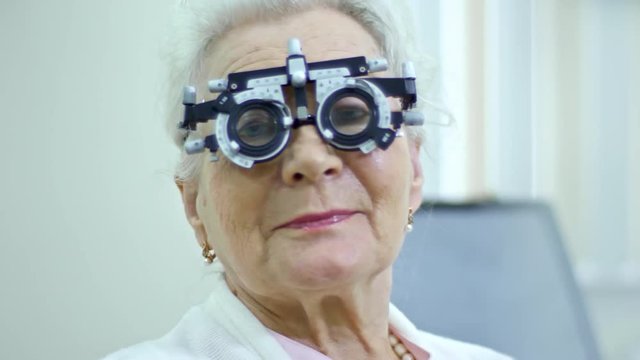 Tracking shot of cheerful elderly woman in trial frame looking at camera while visiting ophthalmologist in eye care clinic; female doctor in background