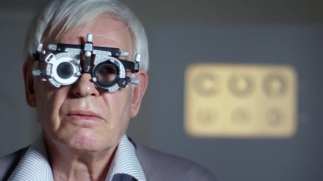 Tracking shot of senior man in trial frame with testing lenses reading eye chart while having eyesight exam at doctors office