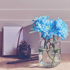 A bouquet of blue chrysanthemums is in a glass jar. Behind there is an open notepad, a black pen, a glass container with a spruce cone inside. Wooden background