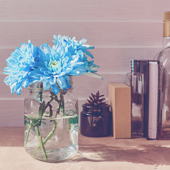 Blue chrysanthemums are in a glass jar. There is a cardboard box, a closed notebook, a bottle with alcohol, a bottle of perfume and a black container with a spruce cone behind it. Wooden background
