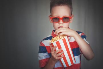 Fototapeta premium Young boy watch a movie in 3D glasses at the cinema or at home. Little kid eat popcorn over gray background. Home theater. Cute Child in vintage cinema eyeglasses