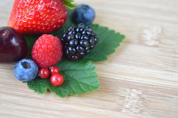Detail of fresh summer fruit and berries on a bamboo board. Strawberries, raspberries, cherries, blackberries, blueberries and red currants on fresh green leaves with copy space.
