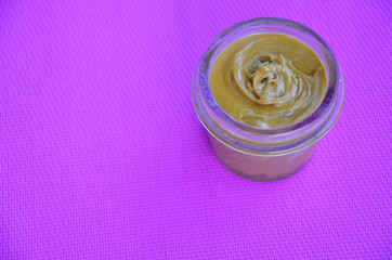 Jar and spoon of peanut butter and peanuts on purple background from top view. copy space