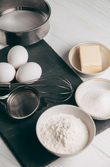 Ingredients and dishes on a white wooden table, which also contains a corolla, are filled with flour, three chicken eggs and a slate board. View from above. Place under the text