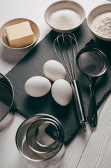 Plakat Ingredients and dishes on a white wooden table, which also contains a corolla, are filled with flour, three chicken eggs and a slate board. View from above. Place under the text