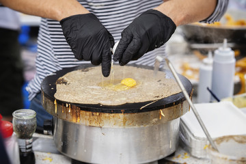 Close-up of hands of cook in gloves preparing crepe galette with ham, cheese and egg on black plate. Concept national food, very tasty fast food, summer party