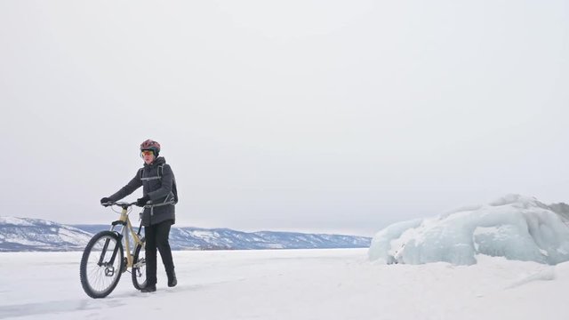 Man is walking beside bicycle near the ice grotto. The rock with ice caves and icicles is very beautiful. The cyclist is dressed in gray down jacket, cycling backpack and helmet. The tires on covered