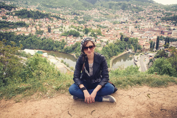 Fototapeta premium Young casual woman sitting on top of a hill, her back turned to the background of the city Sarajevo in Bosnia