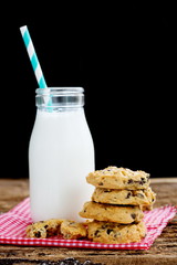 Pile of chocolate chip cookies on napkin with milk in glass bottle.