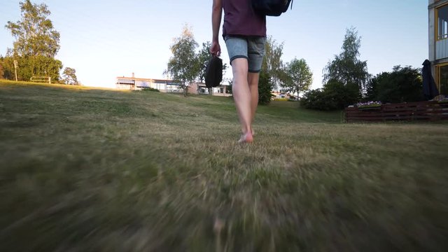 A low Steadicam footage following a Caucasian model walking barefooted in the grass. Beautiful sunny summer evening in Sweden. Barefoot Caucasian male walking outside. Green grass and blue sky.