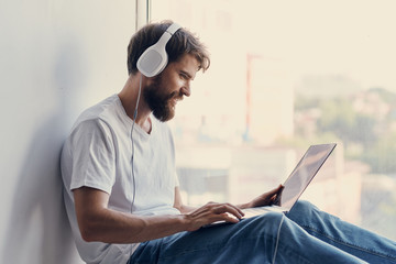 a man with a beard is sitting on the windowsill listening to music with headphones looking at a laptop