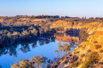 Sandstone eroding cliffs over houseboat moored in Murray River at sunset. Riverland, South Australia