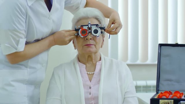 Tracking shot of senior woman sitting in ophthalmology office while female eye doctor trying on trial frame with lenses on her face