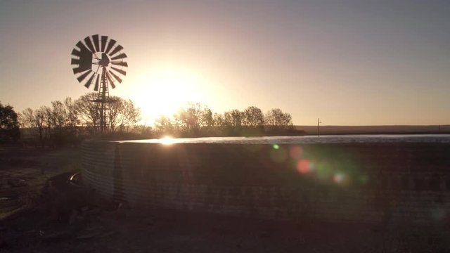 Sun sets with vintage farm wind pump spinning and water storage reservoir.