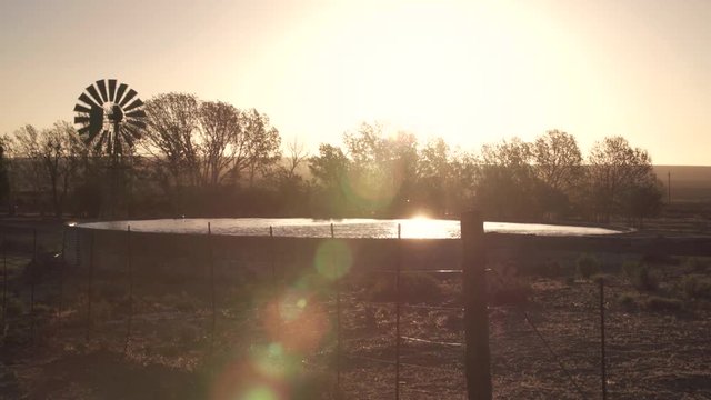 Old fashioned farm windmill spins in the wind and glistening water storage reservoir as the sun begins to set on arid farming landscape.