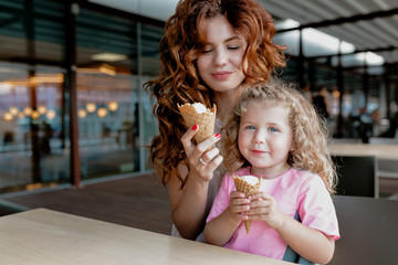Cute little girl and her beautiful mom are eating ice-cream in outdoor cafeteria and smiling while spending free time on thw weekend