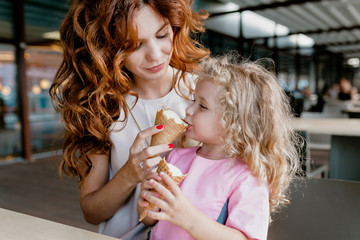 Young pretty woman feeding her daughter ice-cream and having fun while spending weekend outdoor . Portrait of joyful little girl with curly hair having fun with mom in the city