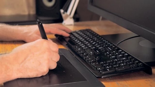 Equipment for graphic design. Touch screen pen, digital tablet, black keyboard and computer monitor. Close up on human hands drawing on tablet computer. Blurred background, soft selective focus