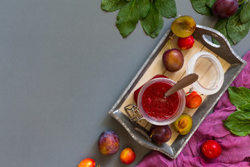 Juicy red plum and plum jam in white utensils and purple runner on gray laconic background pattern with copy space for text flat  lay top view
