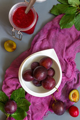 Juicy red plum and plum jam in white utensils and purple runner on gray laconic background pattern with copy space for text flat  lay top view