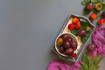 Juicy red plum and plum jam in white utensils and purple runner on gray laconic background pattern with copy space for text flat  lay top view