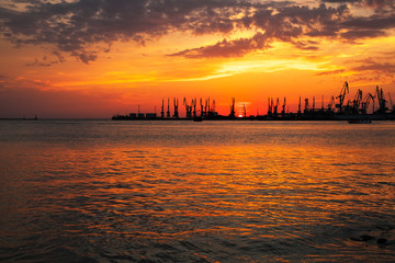 Beautiful landscape with fiery sunset sky and sea. Harbor on the coast during sunrise. Cranes silhouettes against fiery, orange and red sky.