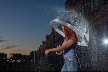 Woman in blue jeans in the rain with transparent umbrella at night. Sad young woman in the rain with umbrella in the evening. Beautiful woman with a transparent umbrella in the lanterns and rain drop