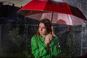 Young woman in green raincoat in rain with red umbrella at night. Sad young woman in the rain with umbrella in the evening. Beautiful woman with red umbrella in lanterns and rain drops