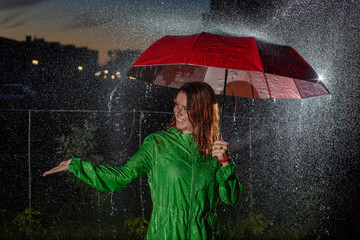 Young woman in green raincoat in rain with red umbrella at night. Smiling young woman in the rain with an umbrella in the evening. Beautiful woman with red umbrella in lanterns and rain drops