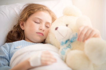 Adorable little girl resting in a hospital bed with her teddy bear. Selective focus at her left eye.