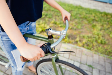 Obraz premium Girl holding the wheel of a bicycle, ready to start riding. Close-up. Dressed in fashionable jeans and t-shirt. Outdoors.