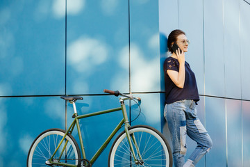 Bottom view to the beautiful stylish girl in sunglasses having a conversation by mobile phone, standing with bicycle at the corner of the modern building